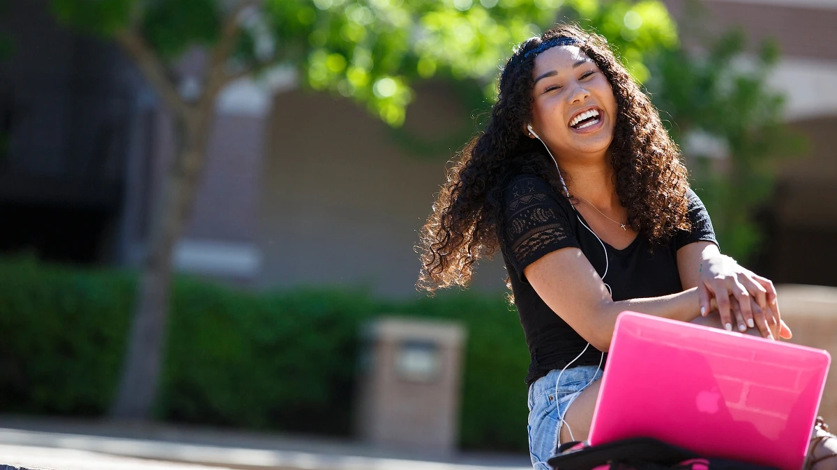 A smiling woman with curly hair is sitting outdoors with a pink laptop and wearing earphones.