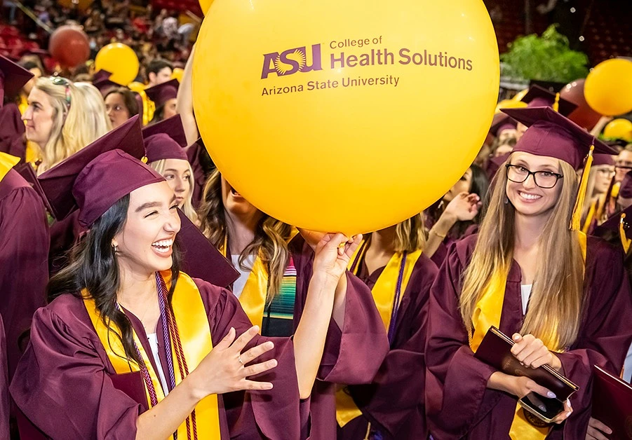 Graduates in maroon caps and gowns smile excitedly as gold balloons with the ASU College of Health Solutions logo drop from above.