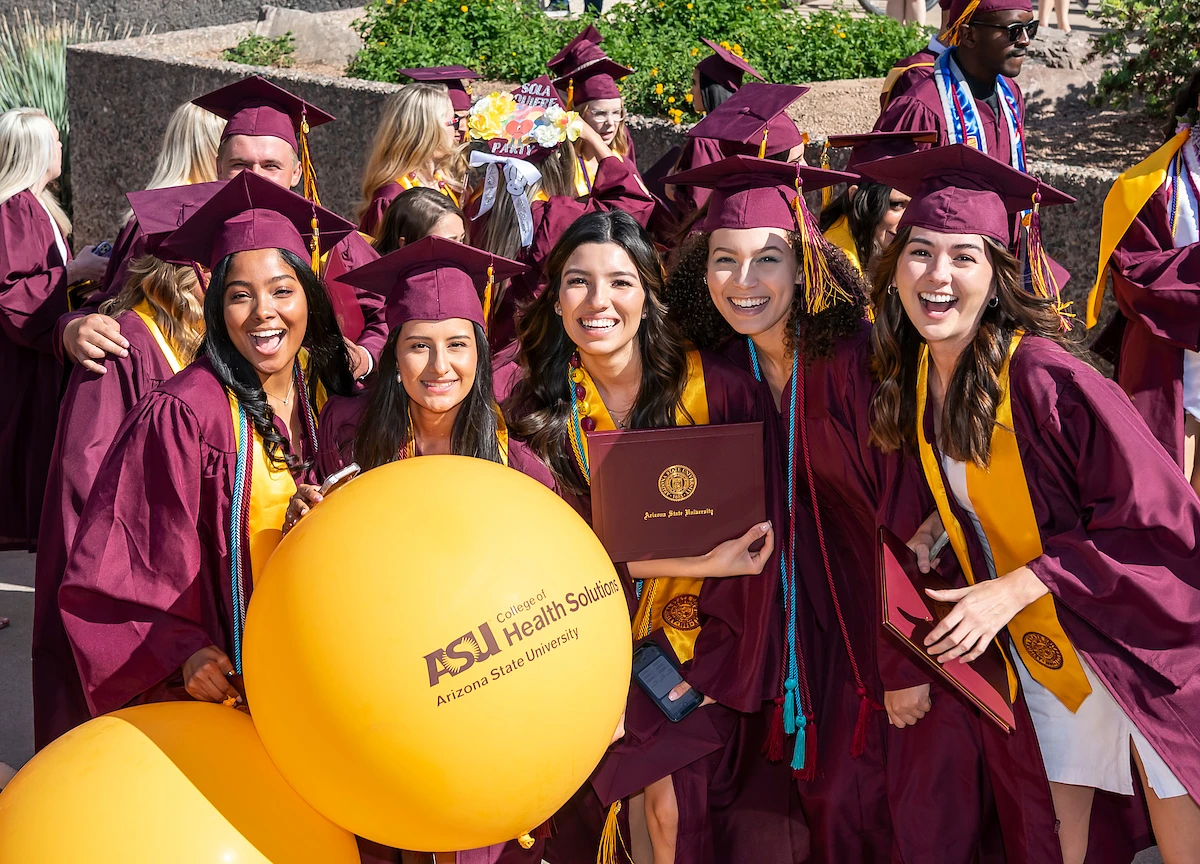 A smiling group of young women in maroon and gold ASU graduation regalia pose for a group photo around a large gold-colored balloon with the ASU College of Health Solutions logo on it.