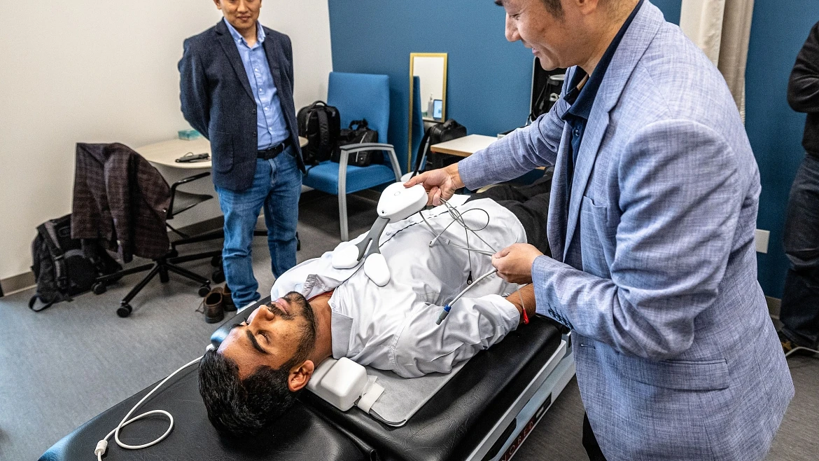 A man in a blue blazer applies a cardiac pressure measuring and monitoring device on a man lying down on an exam table.