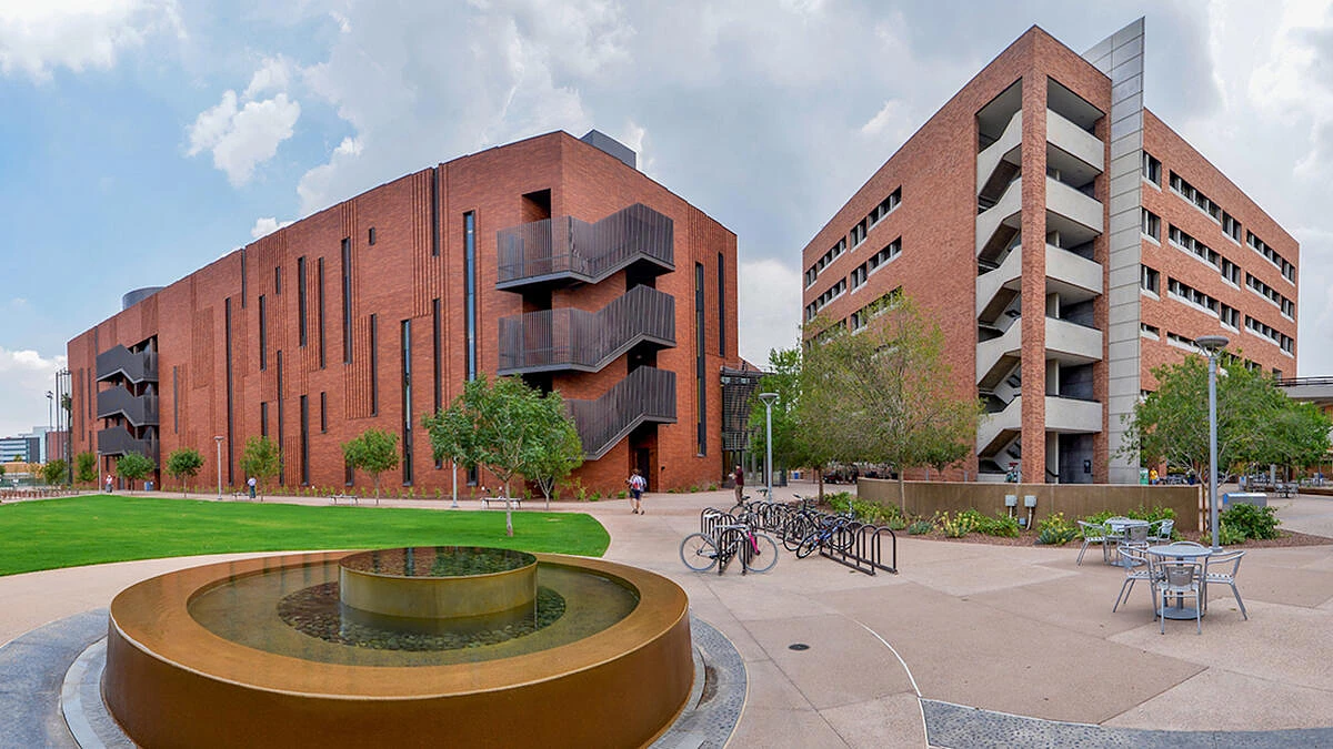 The image depicts two large, modern brick buildings on a university campus, featuring a water fountain, a bicycle rack, and several outdoor seating areas.