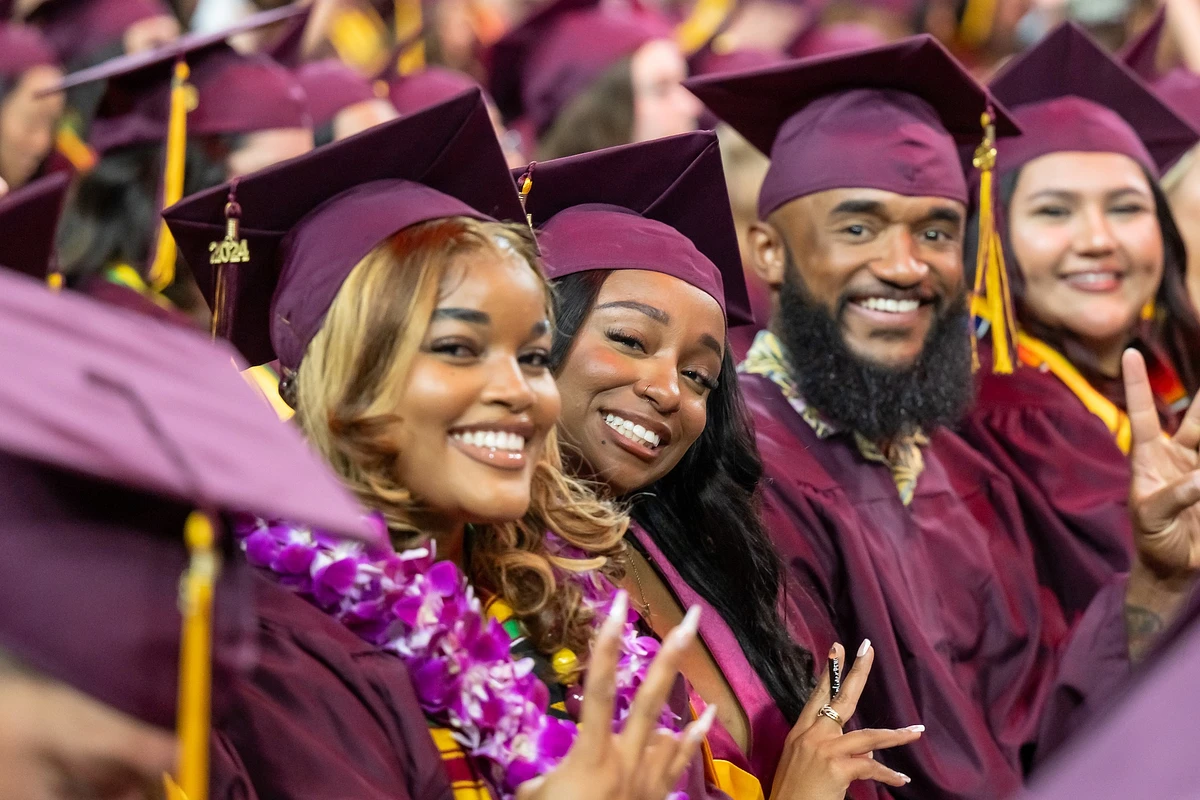 A group of graduates in maroon caps and gowns celebrate with smiles and peace signs.