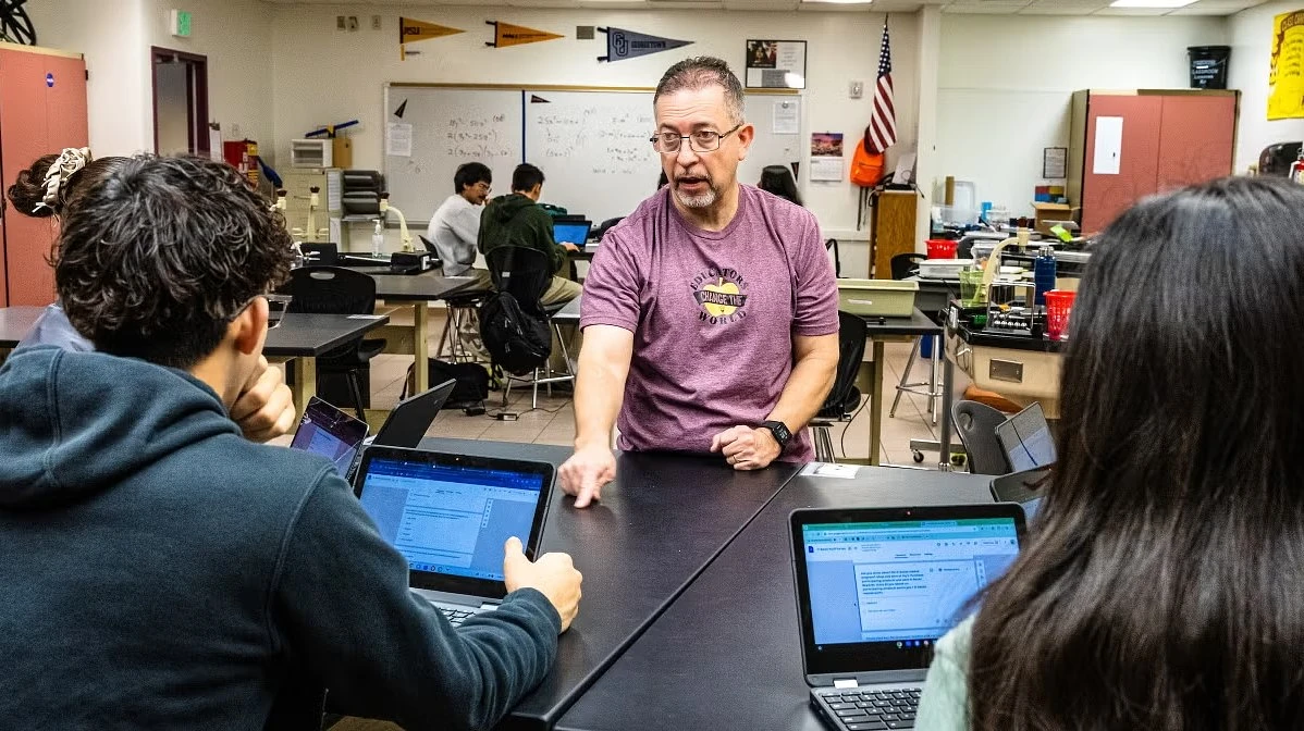 A teacher engages with students working on laptops in a classroom.