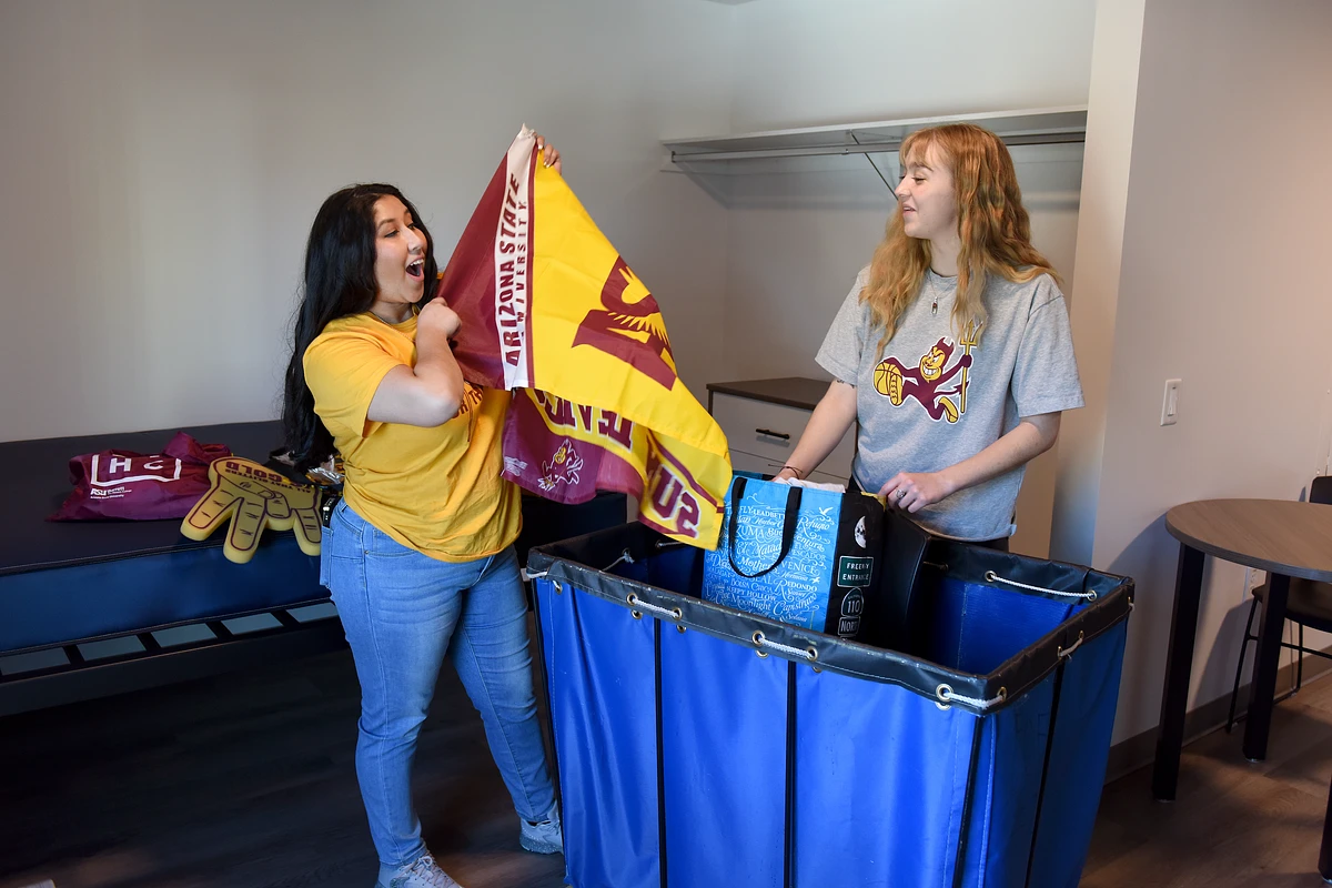Two people are unpacking and holding Arizona State University decorations in a room.