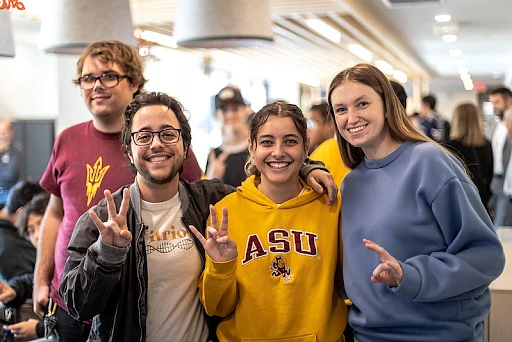 A group of four smiling people are posing together indoors, with two of them making a hand gesture.