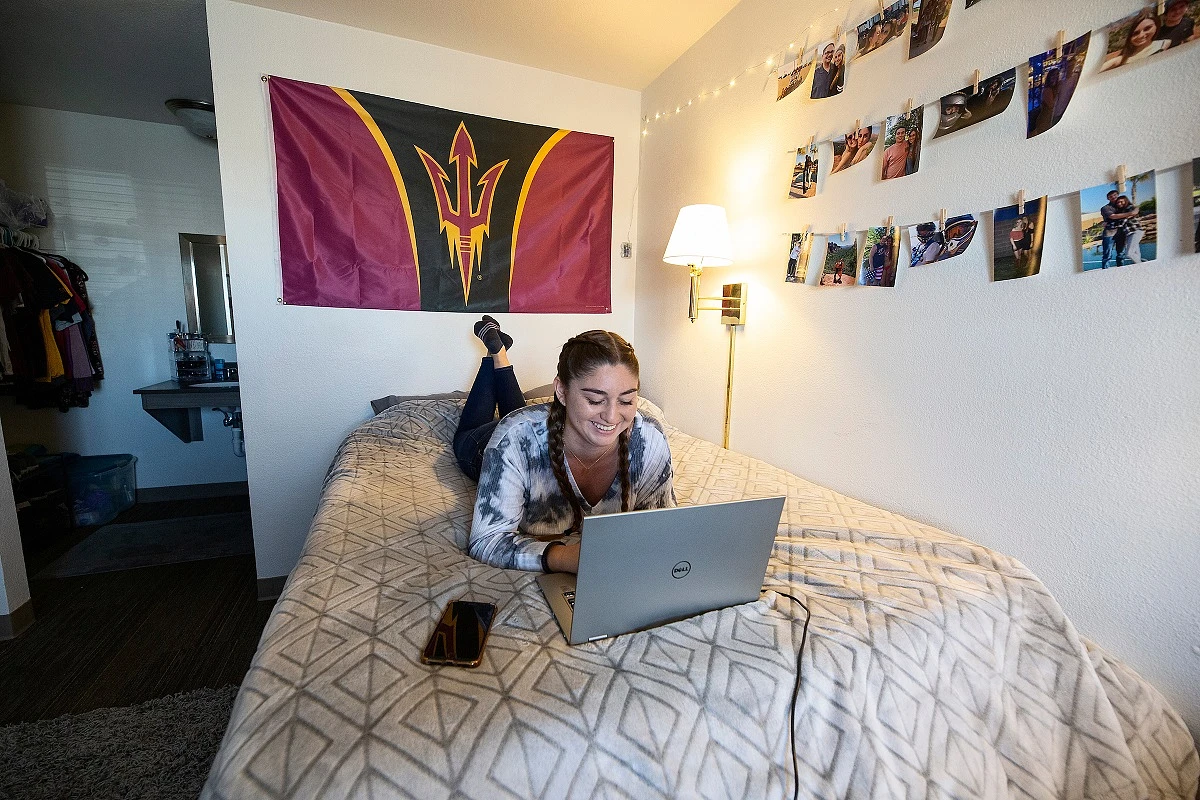 A person is lying on a bed using a laptop in a decorated dorm room, with a college flag on the wall.