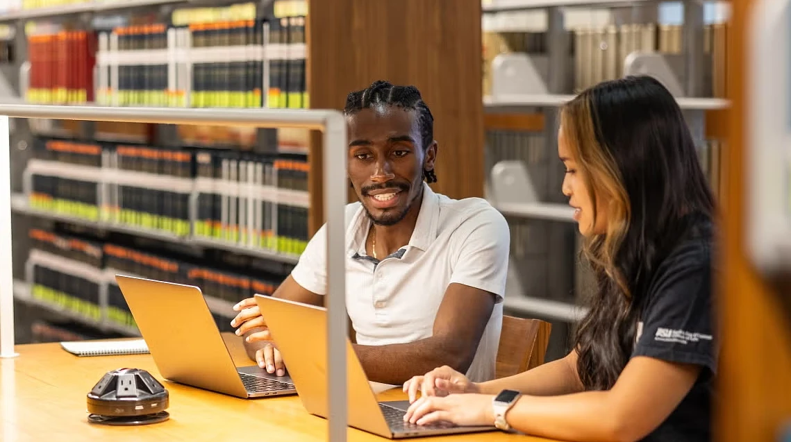 Graduate students working together on laptops in a library setting.