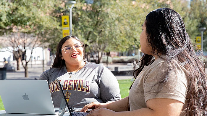 Two people are sitting outdoors with laptops, engaged in conversation and smiling.