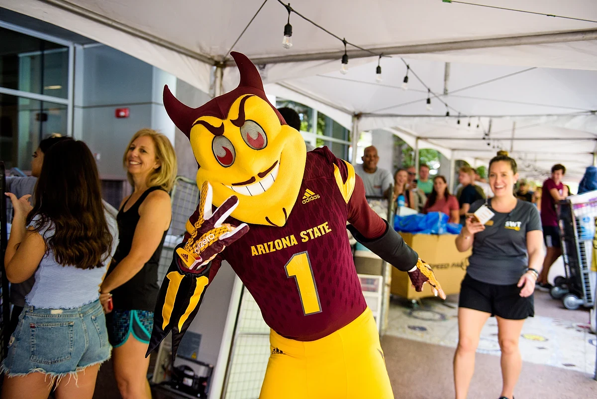 A mascot dressed in an "Arizona State" uniform is playfully posing with a peace sign under a tent, surrounded by smiling people.