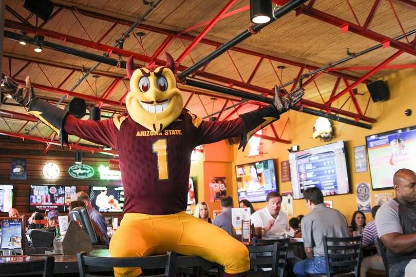 A mascot wearing an Arizona State football jersey enthusiastically poses inside a lively sports bar.
