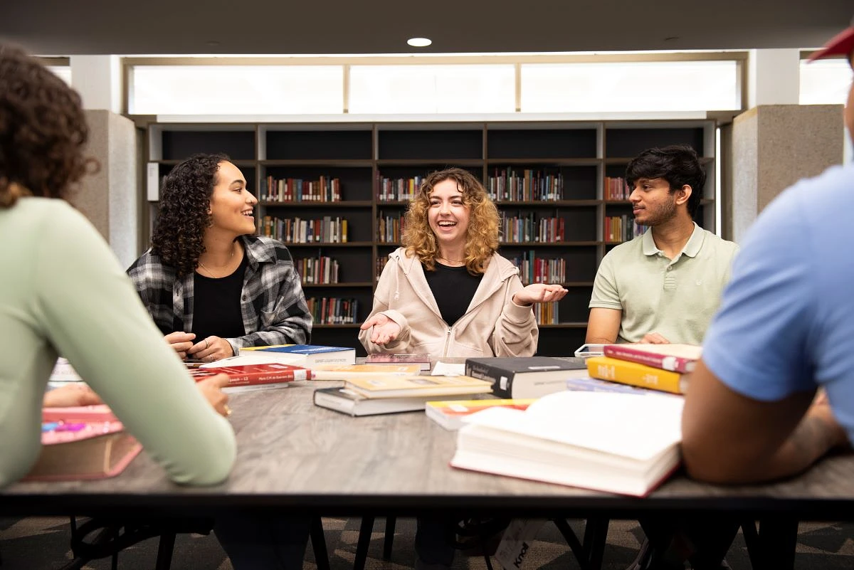 A group of students having a lively discussion around a table filled with books in a library setting.