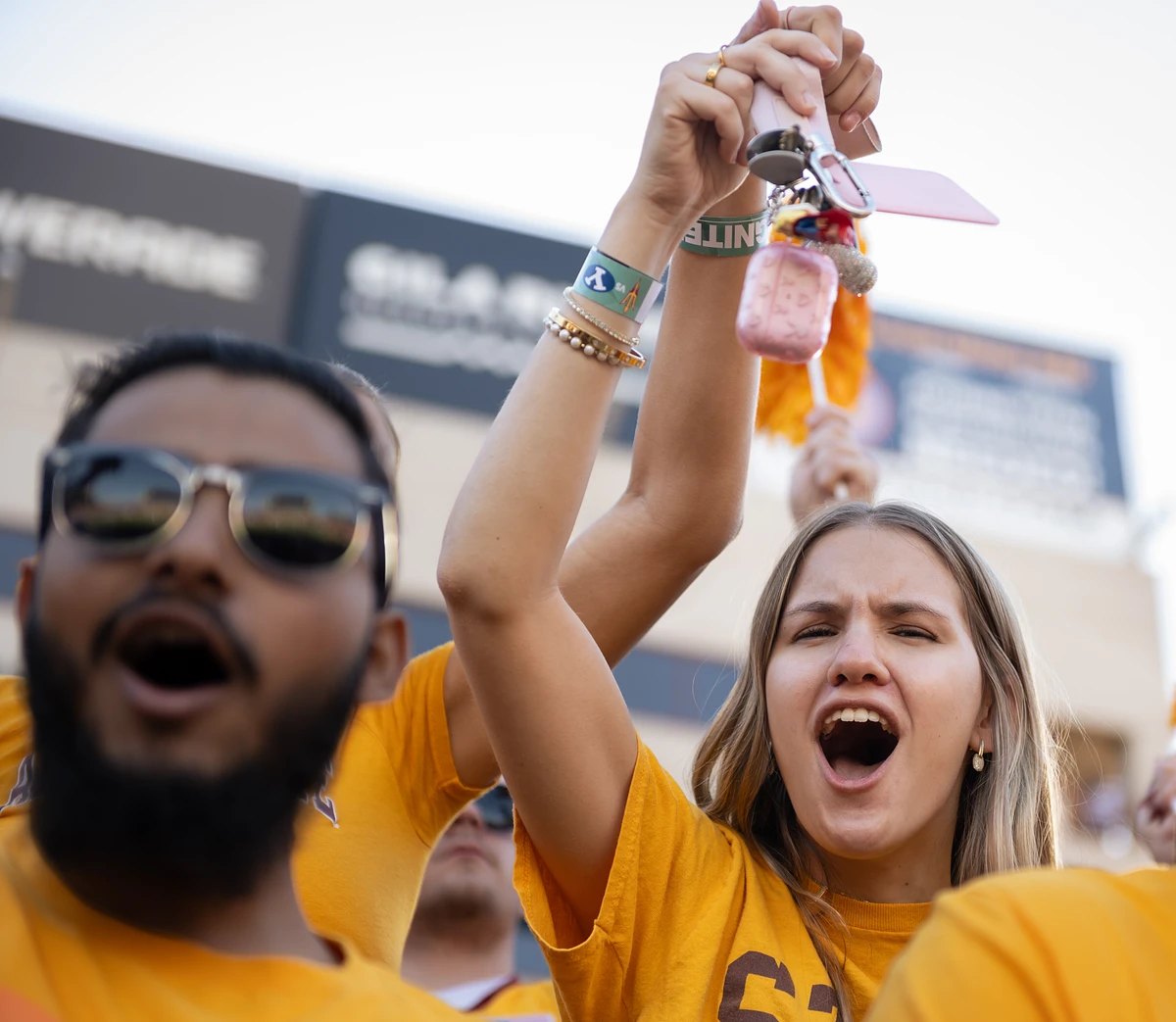 A group of enthusiastic ASU students in gold shirts are loudly cheering at a football game.