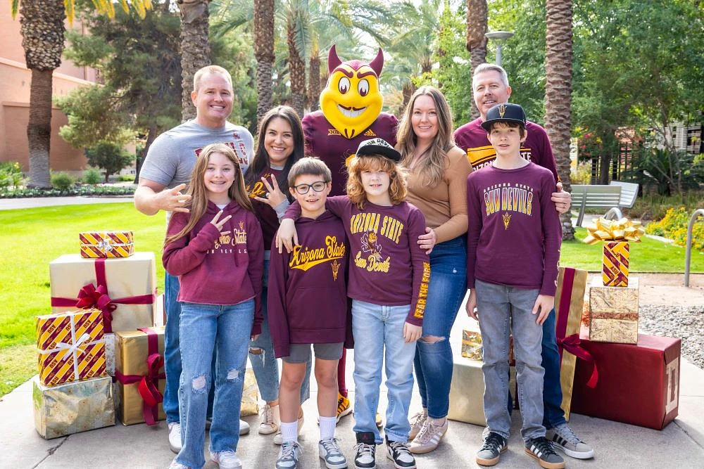 A group of people, including a mascot, pose outdoors with gift boxes, all dressed in Arizona State University apparel.