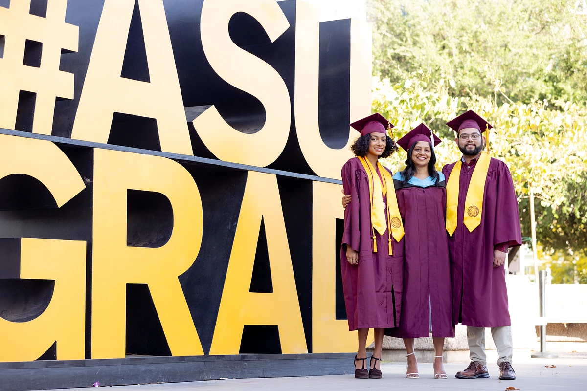 Three graduates in maroon caps and gowns stand smiling in front of a large "#ASUGrad" sign.
